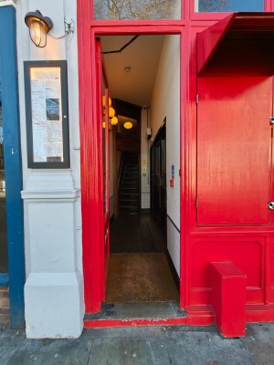 Red wooden entrance door with glass panel ahead. Push from right to open.