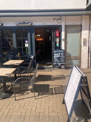 From sidewalk facing venue. Outdoor seating along left. Path ahead to entrance on back. A framed sign on right.
