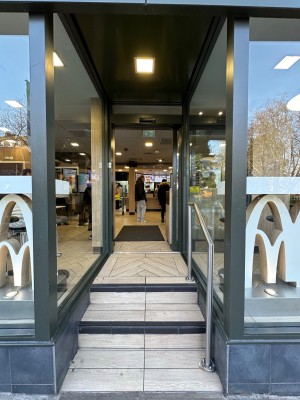 From street, facing venue. Opening ahead. Two steps up to doorway at back. Silver, metal handrail along right.