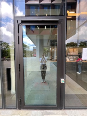 Single glass door with black frame. Push green button on its right to open. Door opens outwards.