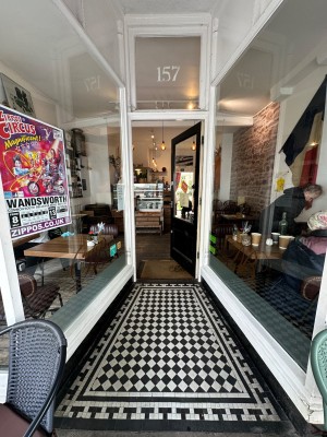 From street, facing venue. Glass venue walls on both sides. Glass door with black, wooden frame at back. Push silver handle on left to open.