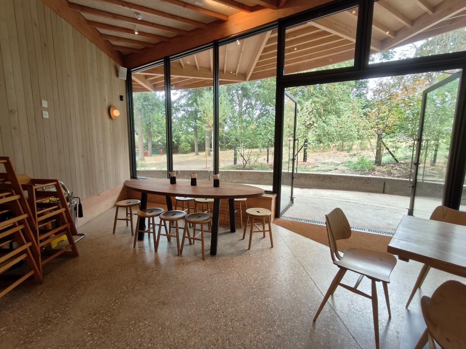 Stacked chairs along left wall. Tables and stools in centre. Tables and open doors on right.