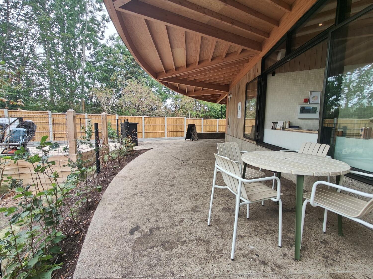 Planting beds along left. Paved path curves ahead. Tables and chairs on right.
