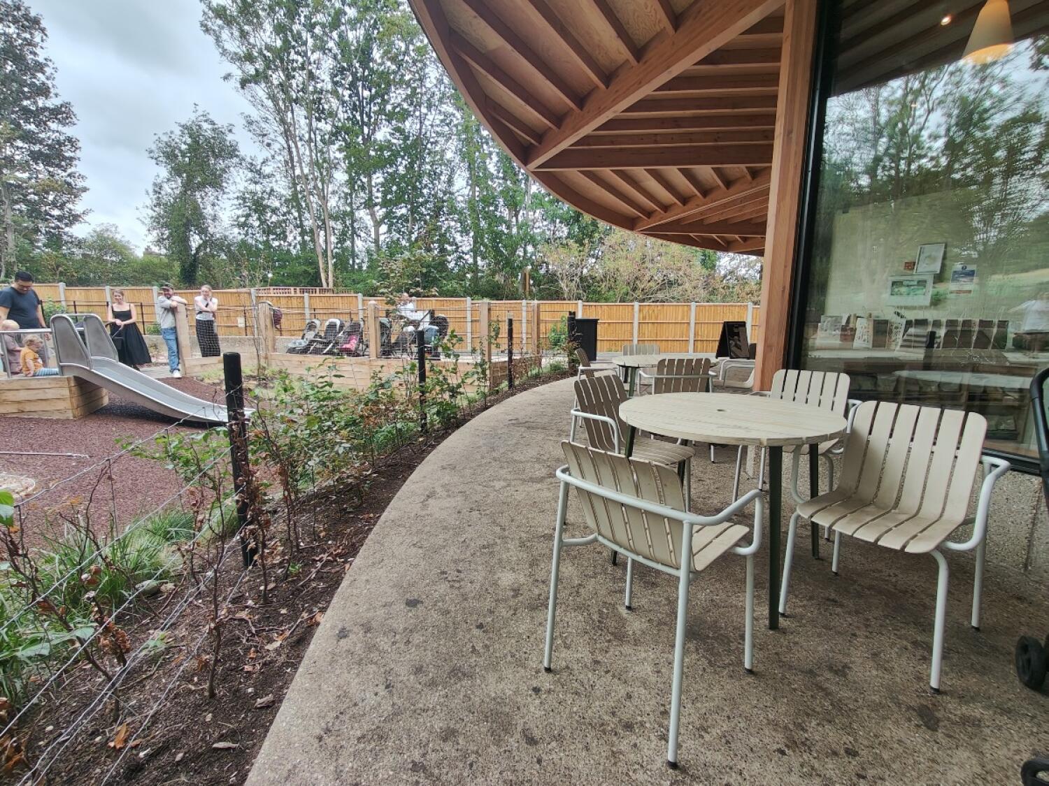 Play area and planting left. Curved concrete path in centre. Tables and chairs along right.