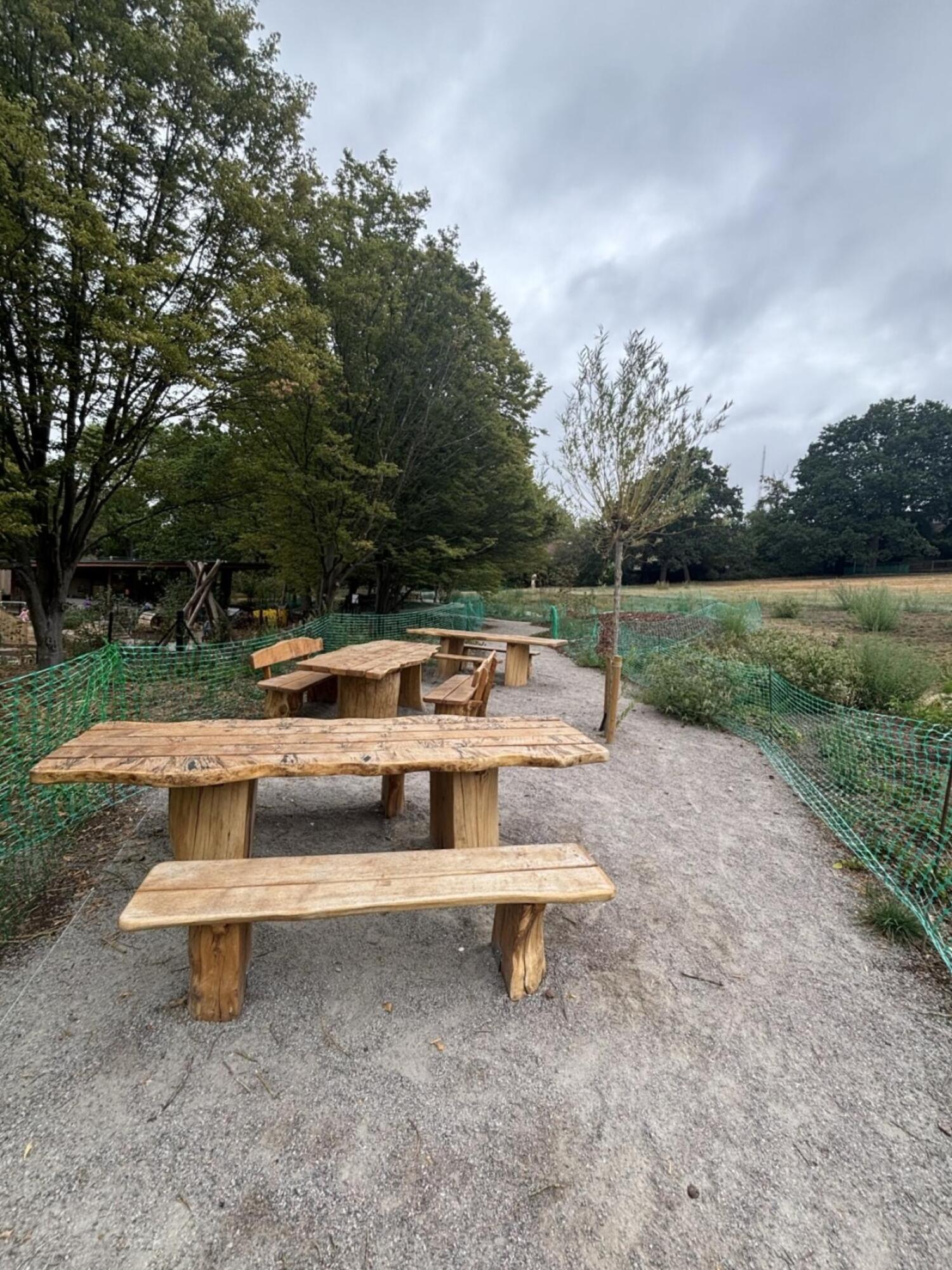 From end of path facing straight. Picnic tables and benches along left and back. Fencing on both sides of path.