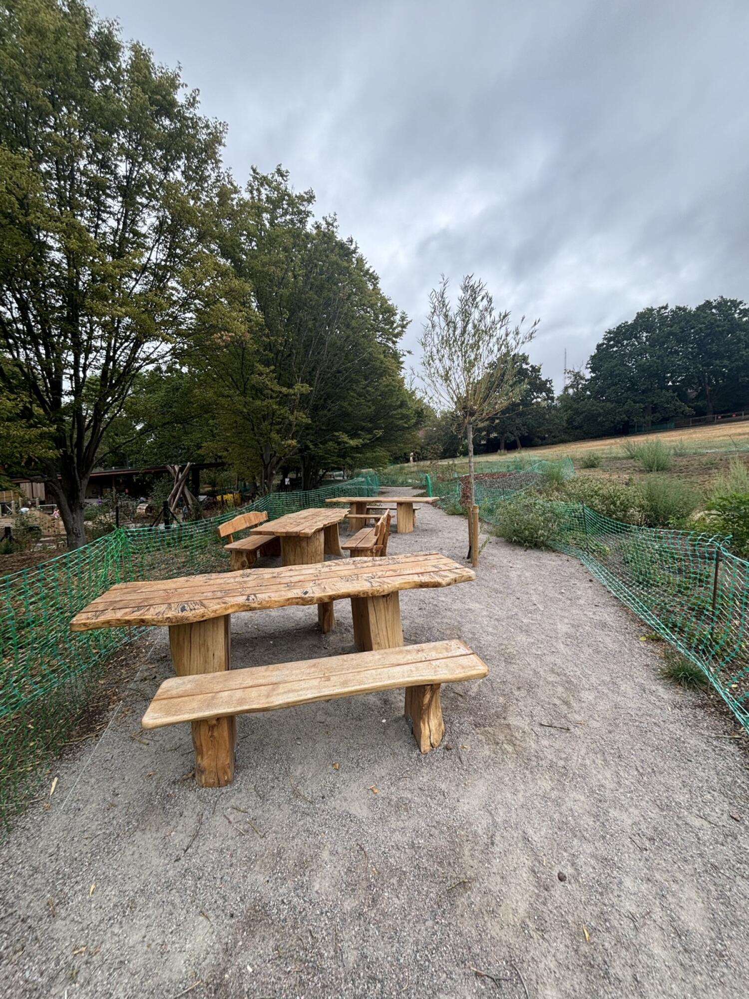 Picnic tables along left. Gravel path continues ahead. Green mesh fencing both sides.