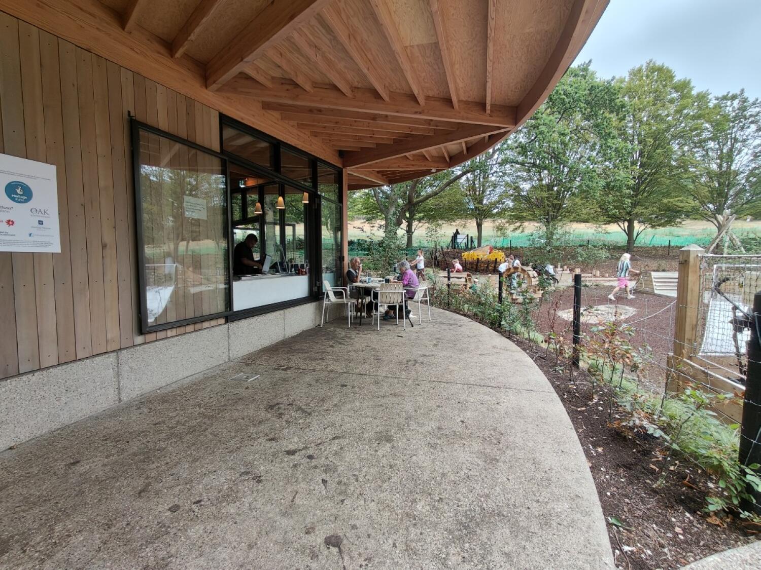 Building and service window left. Paved path with tables centre. Fenced garden on right.
