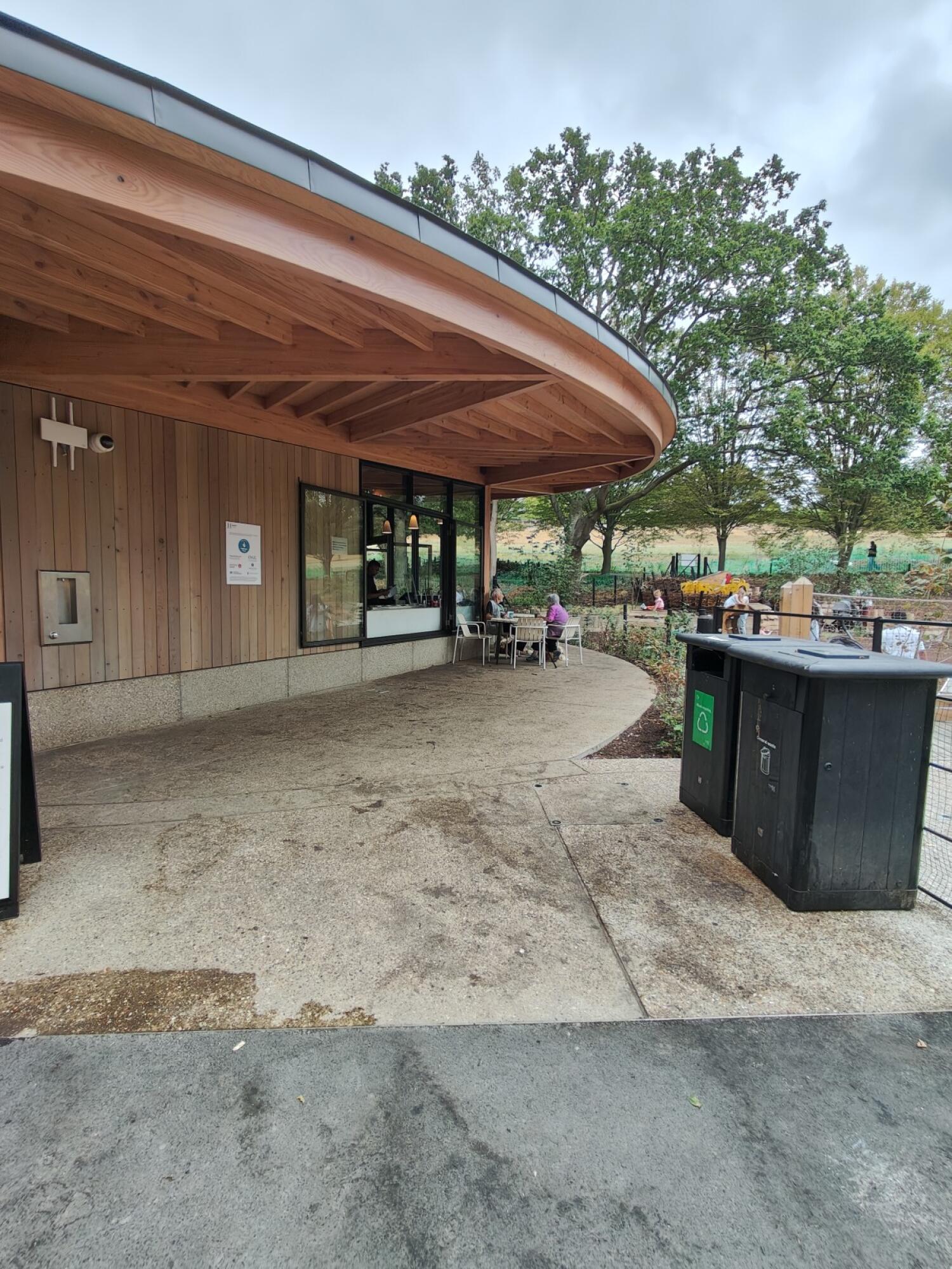 Paved path beside building. Outdoor tables in centre. Bins on right.