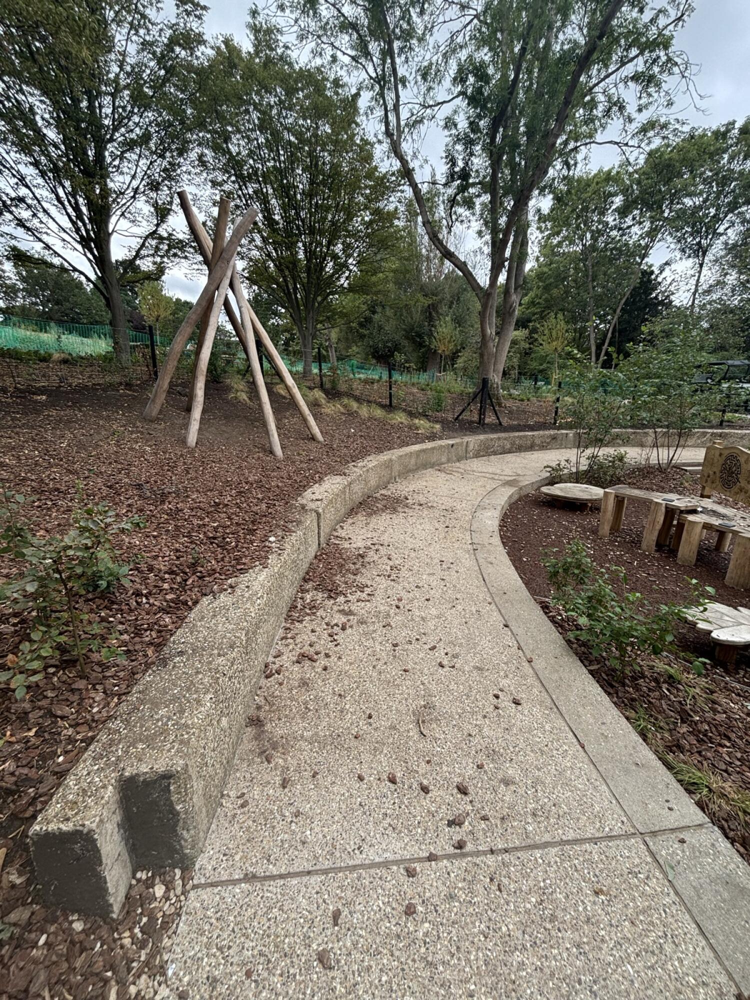 From after opening facing straight. Curved path ahead through garden area. Fixed benches along left of path.