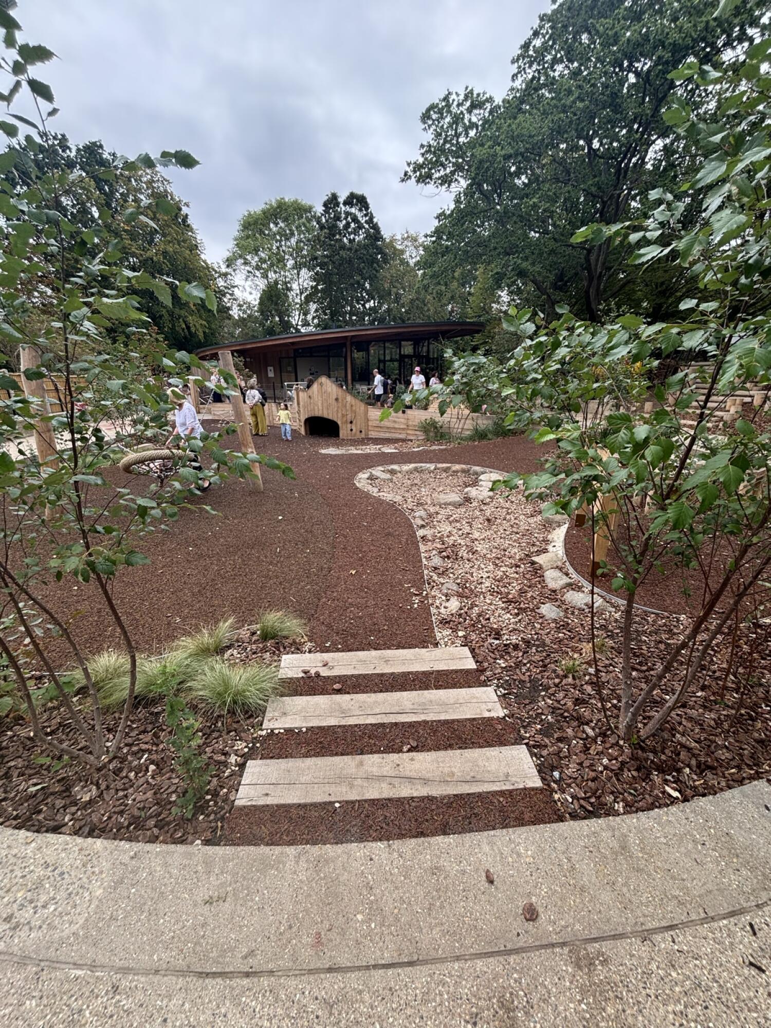From after curved path facing right. Three steps ahead to play equipment on at back. Plant beds on left and right of steps.