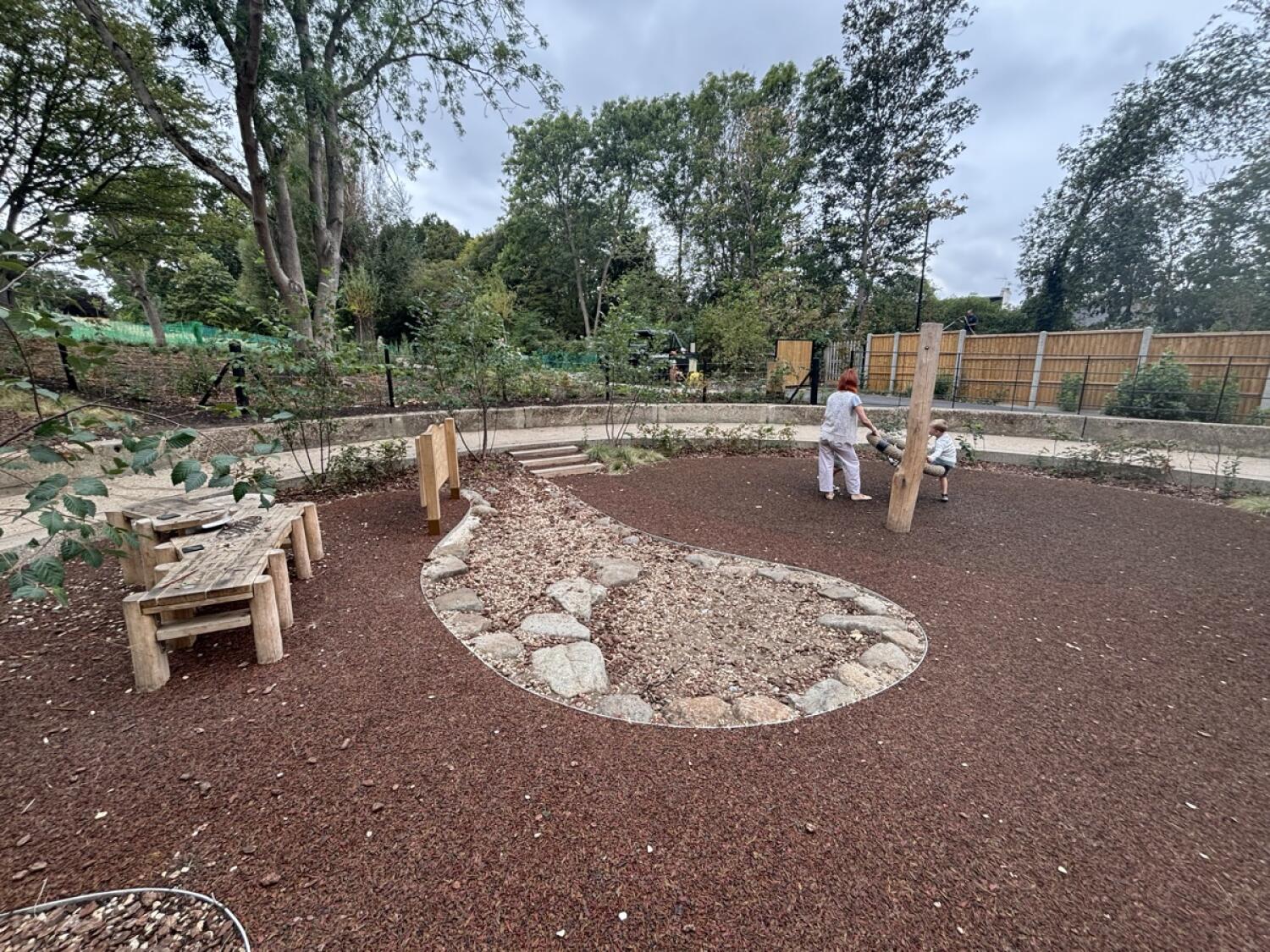 From after plant beds facing straight. Wooden play tables on left. Gravel play area in centre.