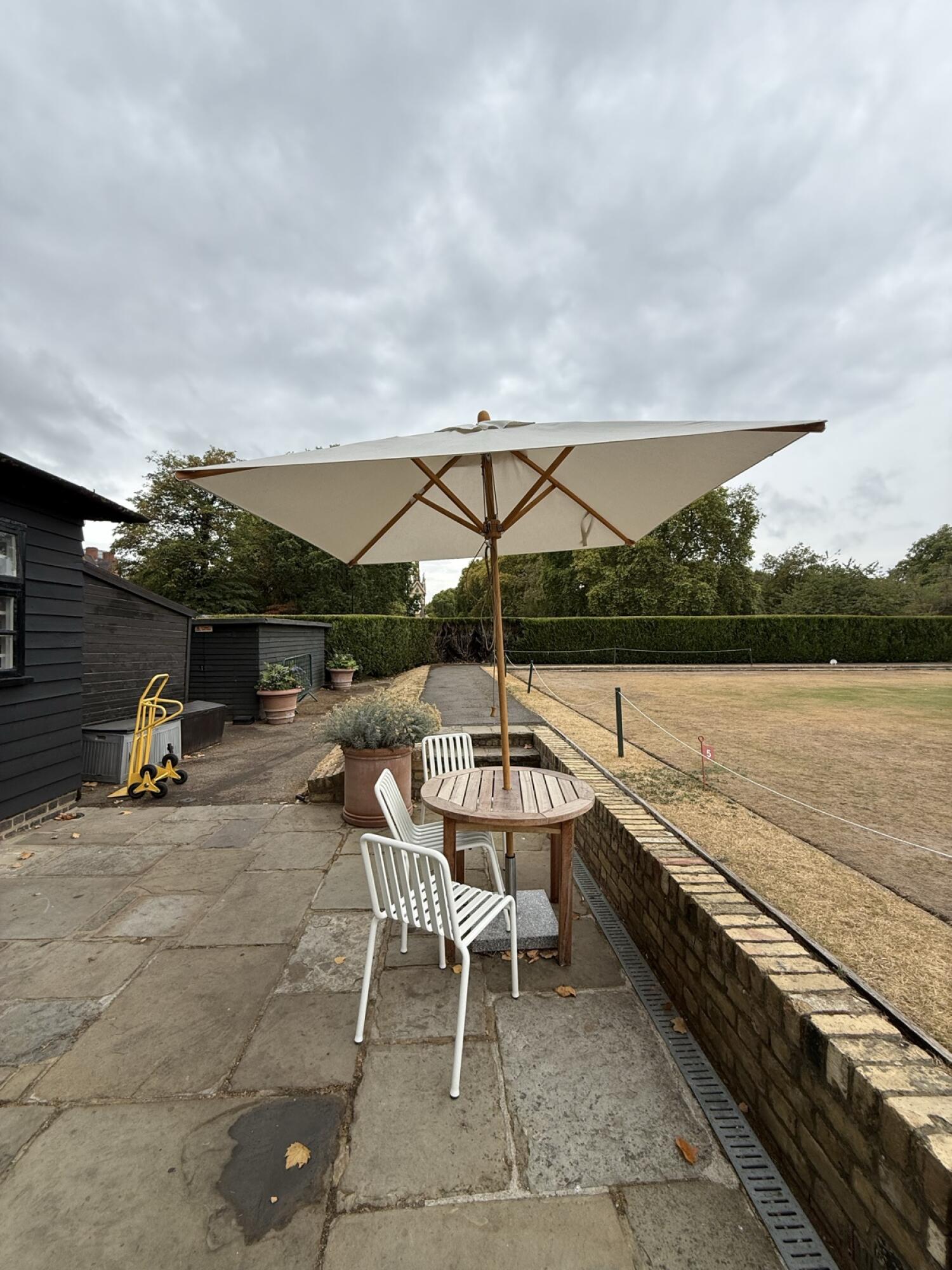 From bottom of ramp facing back. Table, chairs, and parasol centre. Low wall and lawn right.