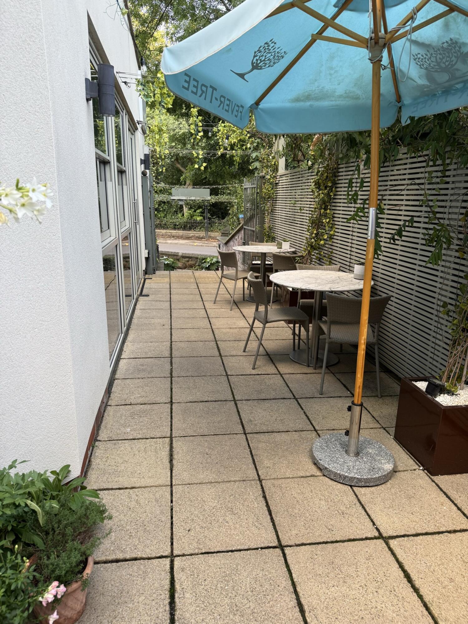 Glass doors along left. Paved path through centre. Tables, chairs and parasol right.