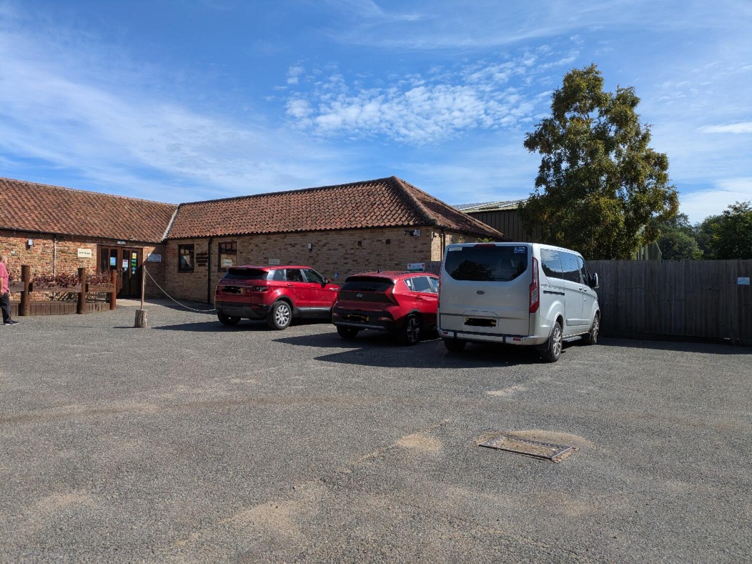 Entrance and seating on left. Gravel car park with parked vehicles. Fence along right.