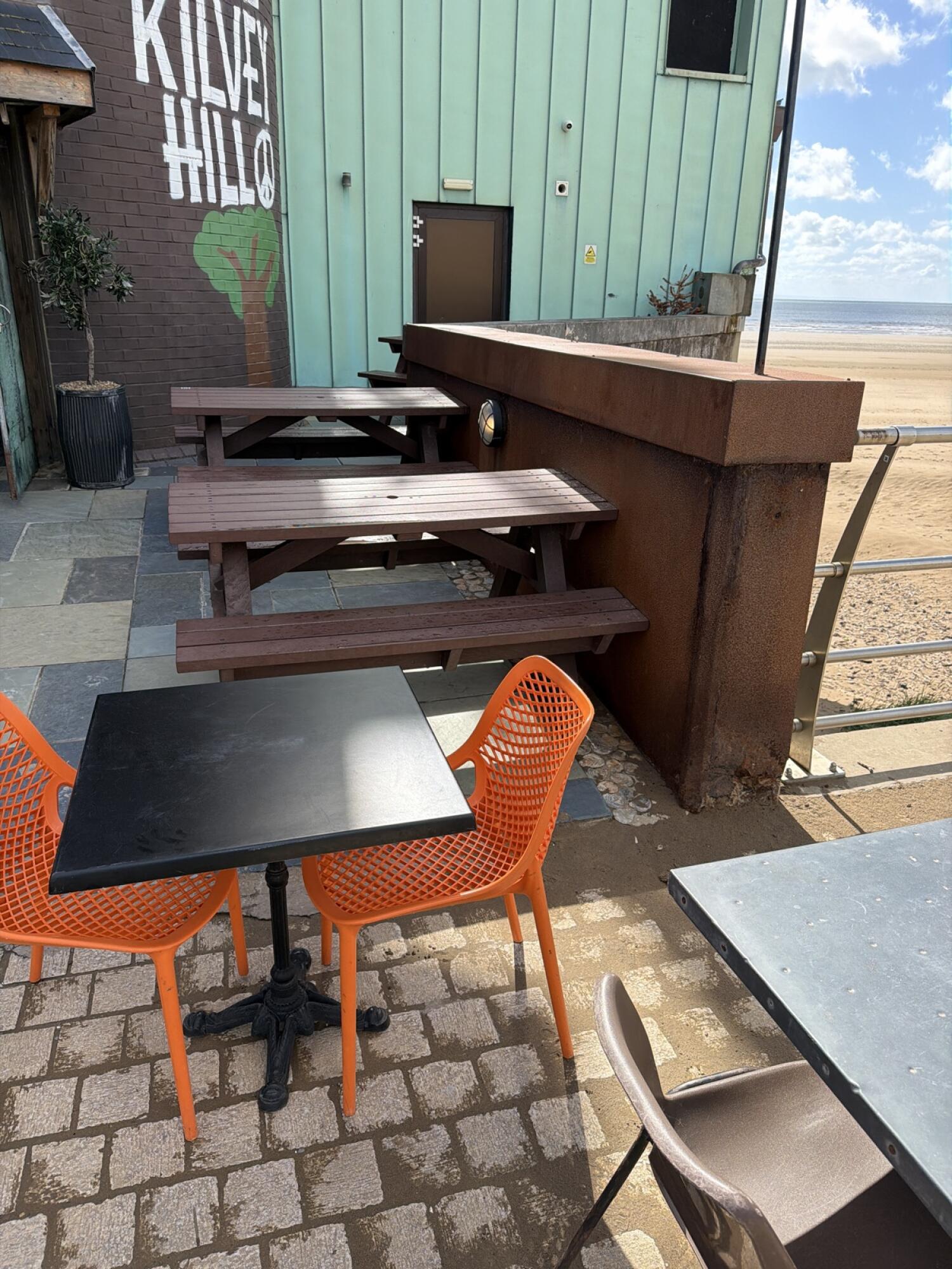 Picnic benches along left wall. Tables and chairs in centre. Railing right with sandy edge.
