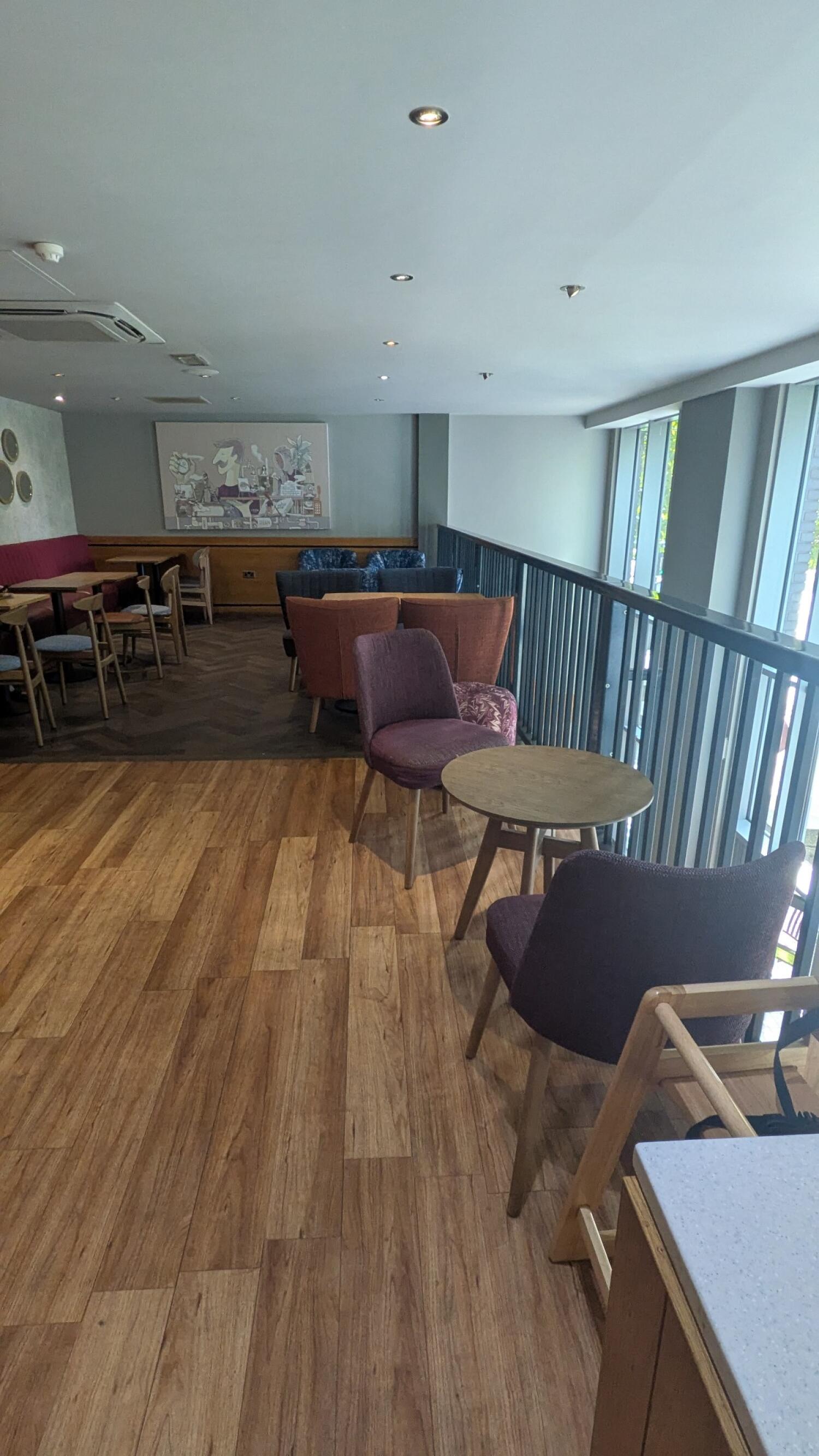 Tables and chairs along left. Armchairs on darker flooring centre. Railing and windows along right.