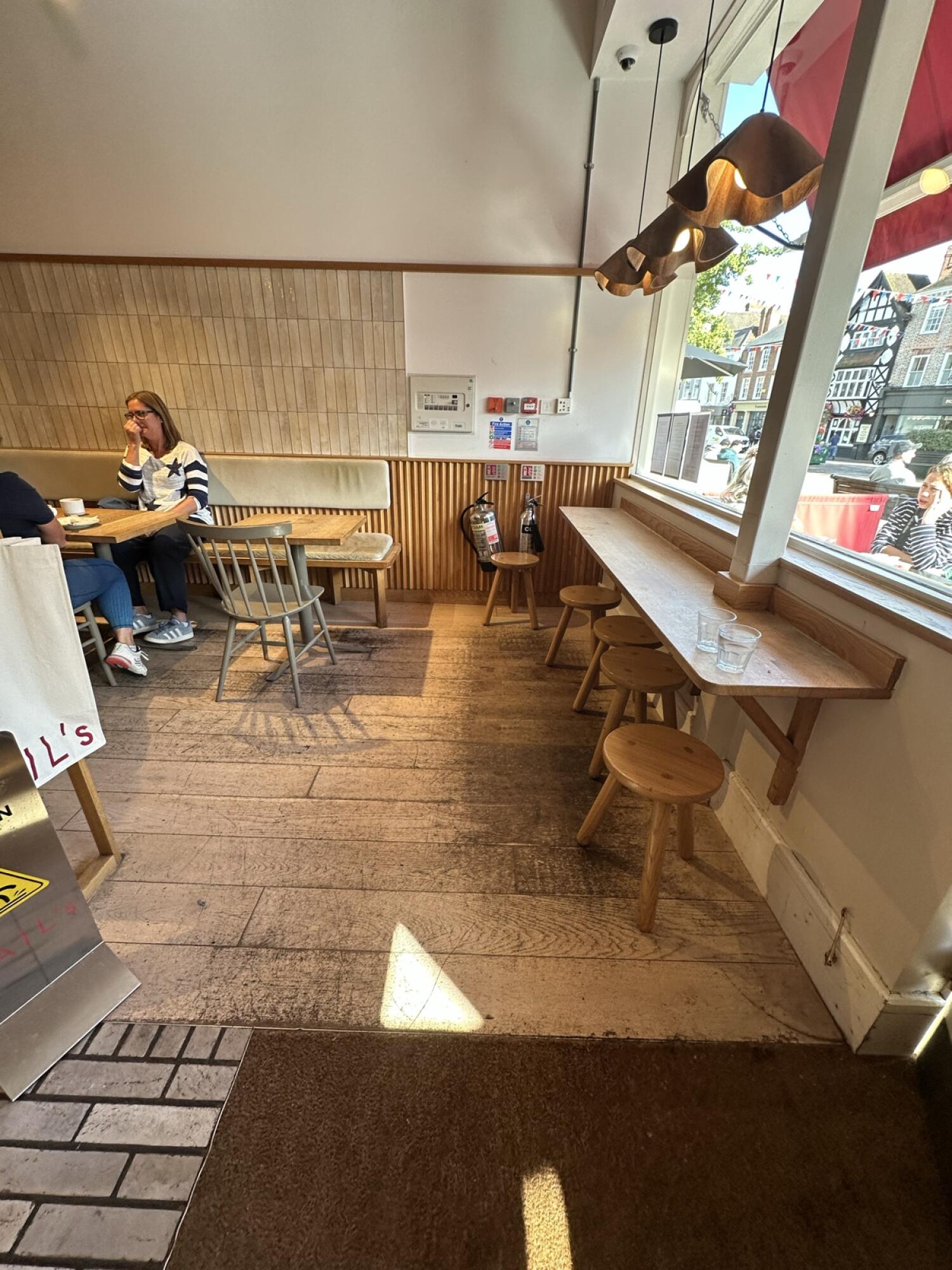 Tables and chairs along left. Wooden floor space in centre. Window counter with stools right.