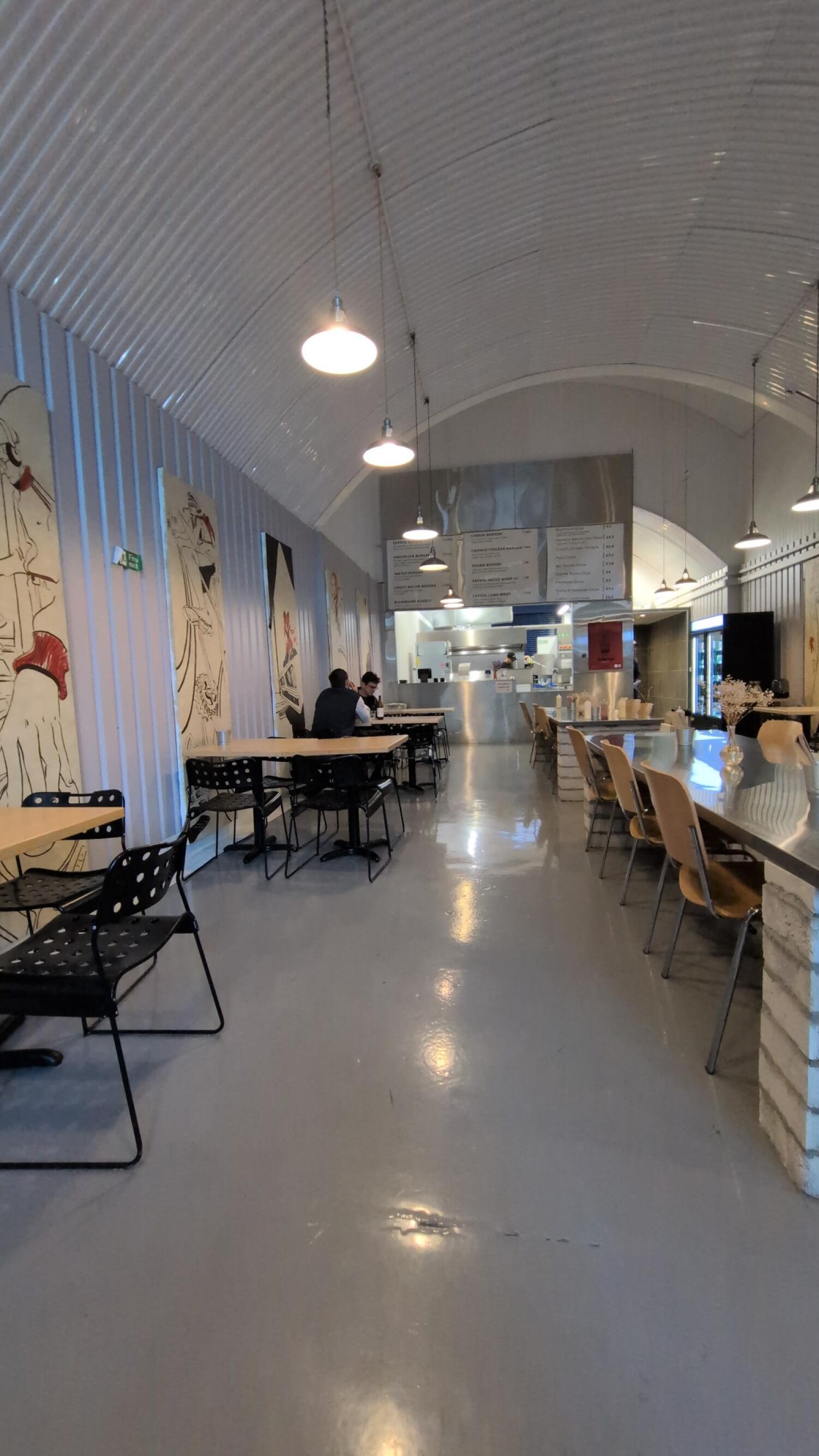 Tables and chairs along left. Path to service counter centre. Counter seating and fridge right.