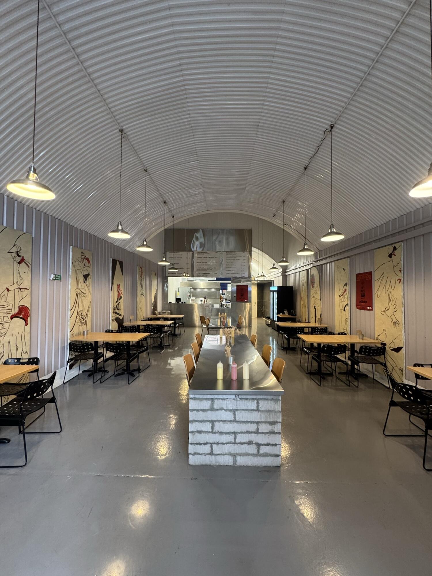 Tables and chairs along left. Long table centre, service counter behind. Tables and chairs along right.