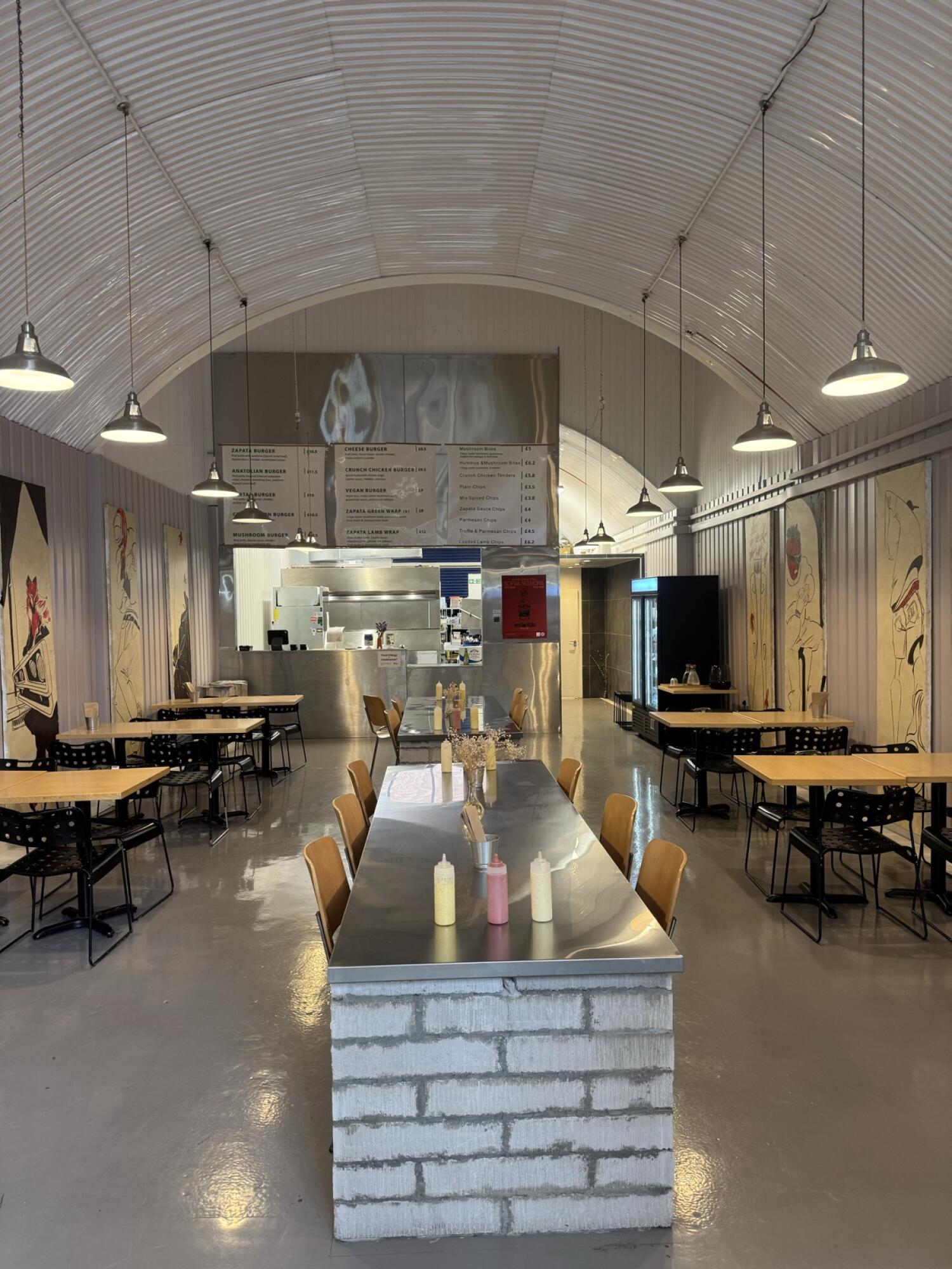 Tables and chairs along left. Communal table centre, counter behind. Fridge and seating along right.