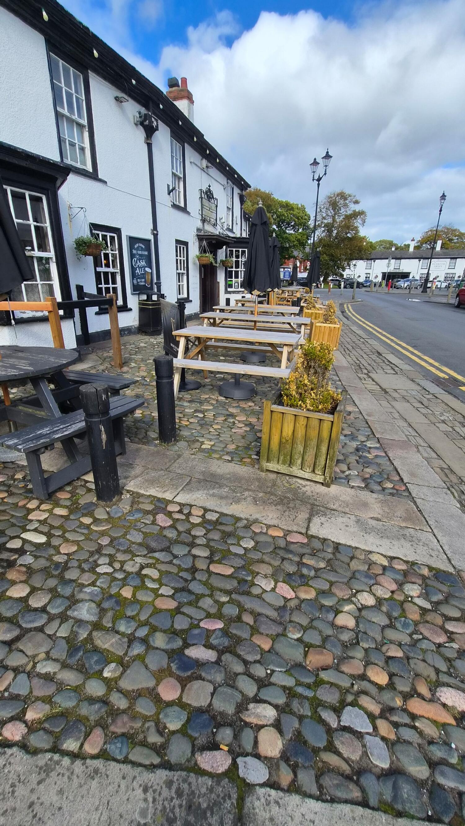 Pub building along left. Picnic tables on cobblestones centre. Planters border pavement on right.
