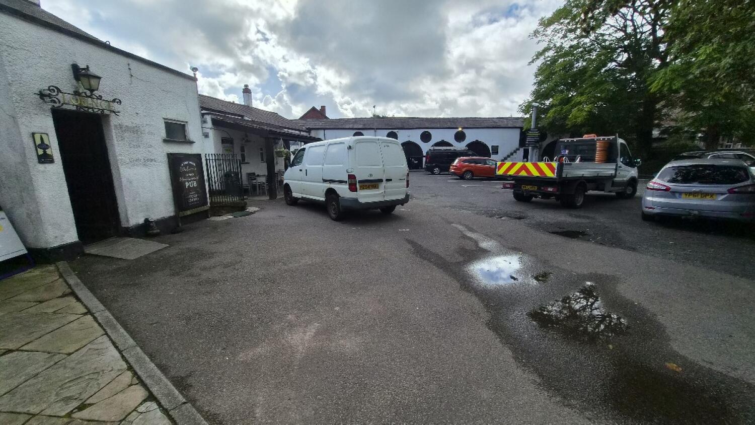 Pub entrance and sign on left. Car park space in centre. Puddles and parked vehicles right.
