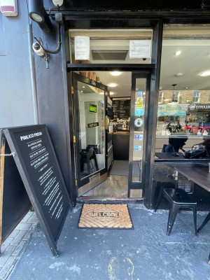 Brown doormat immediately before door. Wooden step at threshold of door. Black wood-framed glass door. No handle. Push on right side to open.