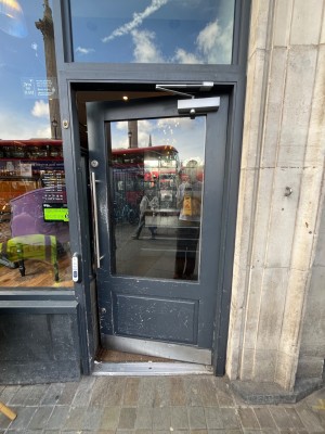 Grey framed glass door ahead. Push open via long silver handle on left. Doorbell for assistance left of door.