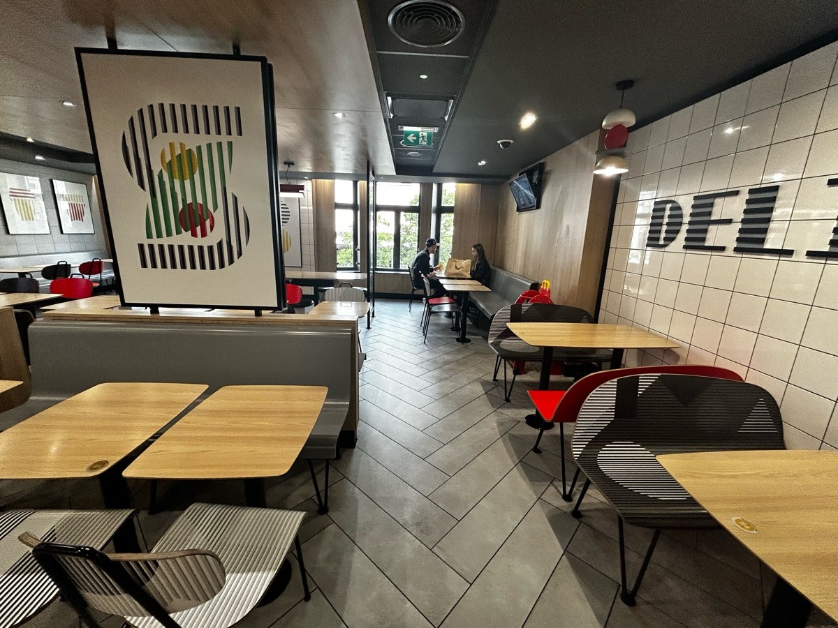 From end of long table, facing straight. Half booths on left. Path ahead leading to half booths on  corner of back wall. Chairs and tables on right