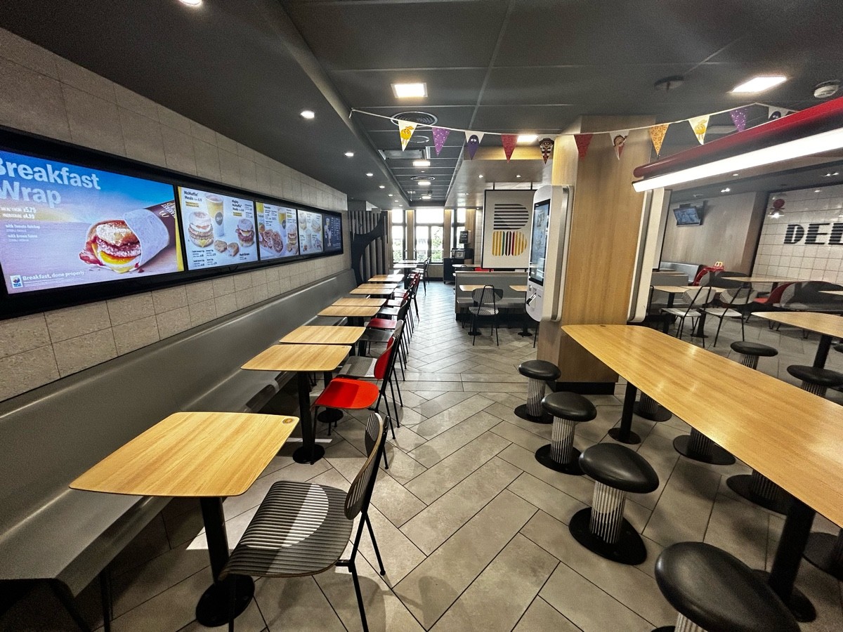 From preceding door, facing left. Half booths along left. Path ahead. Stools and long table on right followed by pillar with online ordering machine