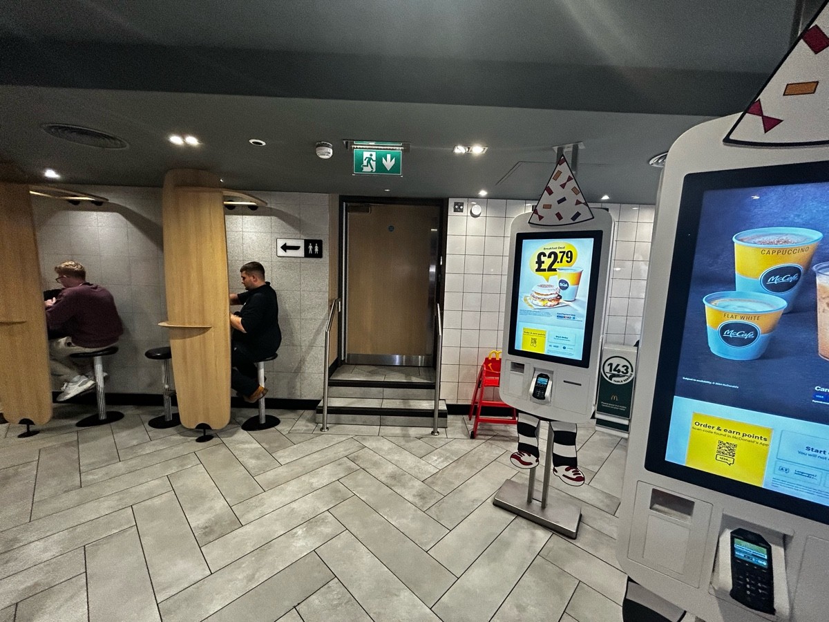 From preceding door, facing straight. Online order machines on right. Path ahead leading to stairs and private area. Panels covering stools on left.