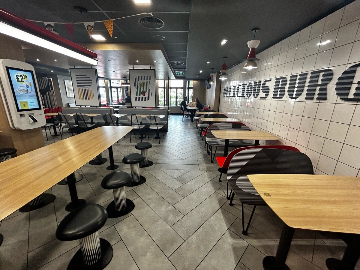 From end of path, facing left. Long tables with fixed stools on left. Path ahead. Tables and chairs on right along wall.