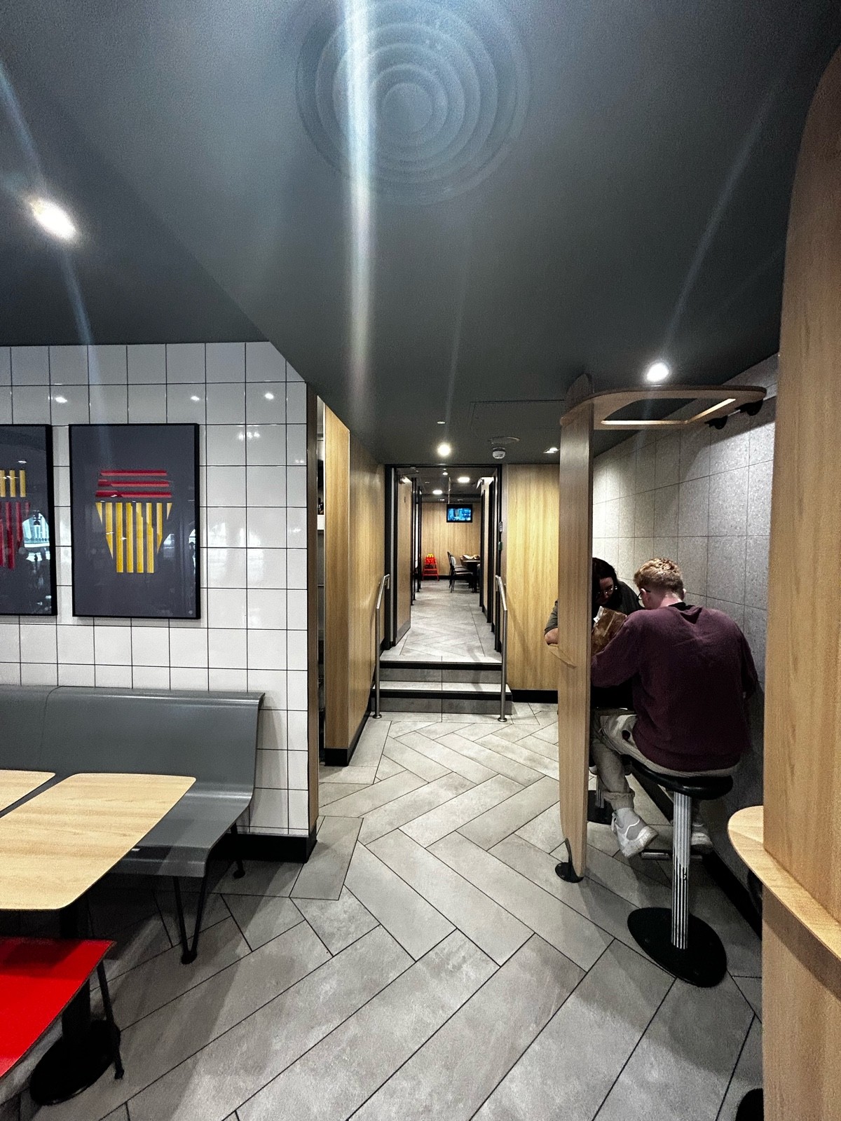 From panels, facing left. Half booth on left. Path ahead leading to stairs and seating area. Stools and wood panels on right.