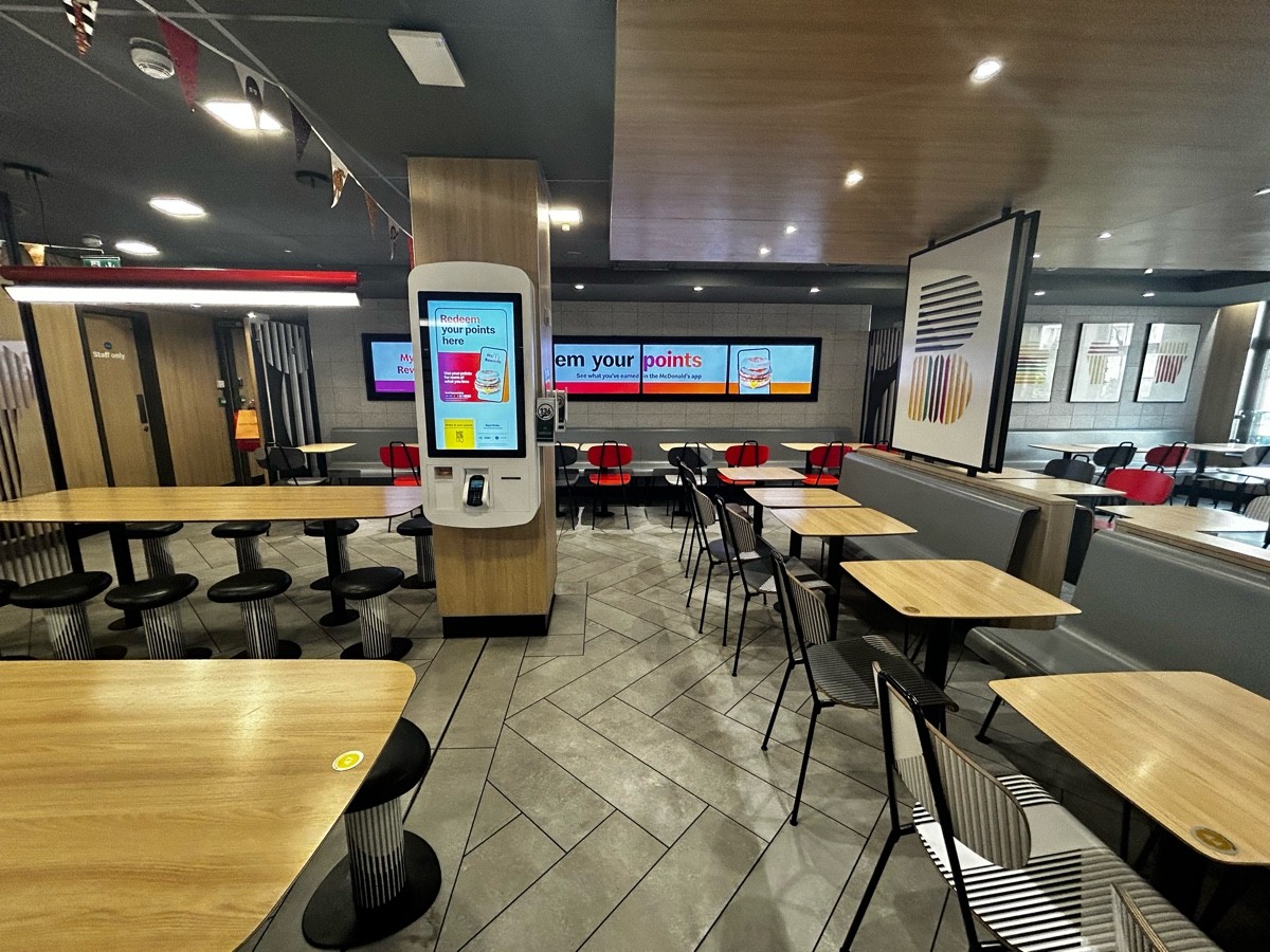 From end of long table, facing left. Stools and long tables on left. Path ahead obstructed by pillar with online order machine. Half booths on right
