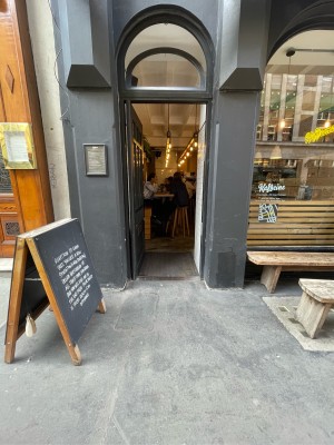 Dark grey and glass panelled door ahead. Push brass handle on right to open. A-board on left, benches on right.