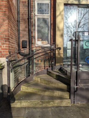 From pavement facing stairs. Curved cement stairs ahead with metal handrail and glass wall along left. Entrance at top of stairs.