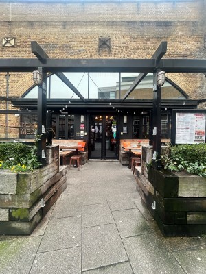 From street facing venue. Planters to left and right. Archway ahead. Outdoor seating to left and right. Entrance at back.