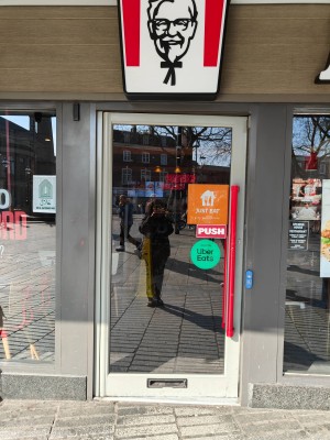 Glass door with grey frame ahead. Push red handle on right to open. Doorbell to right of door.