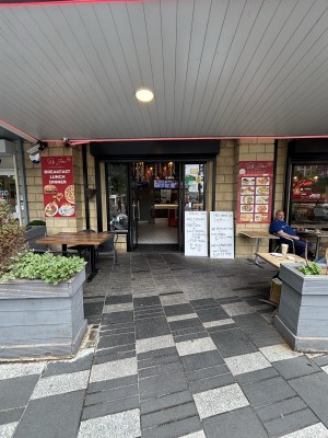 From sidewalk facing venue. Opening ahead between planters. Path behind between outdoors seating to doors on back.