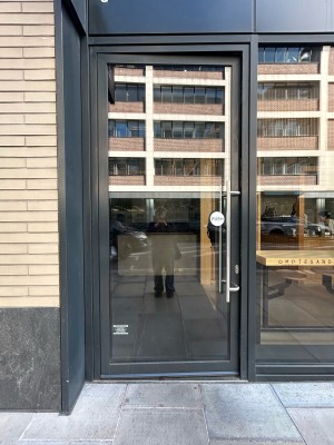 Single glass door with dark grey frame. Push vertical silver metal handle along right to open.
