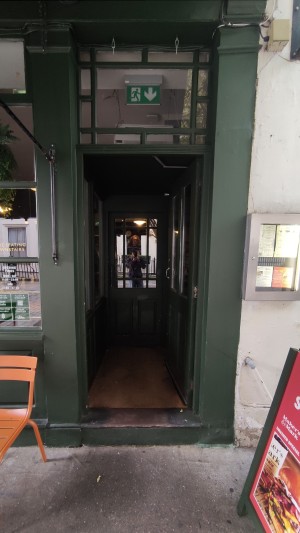 From sidewalk facing venue. One step up to glass door with green frames. Push vertical metal handle to left to open.