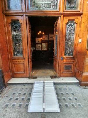 Facing wooden-framed double glass doors. Push golden metal handles in centre to open. Ramp on request before the door.