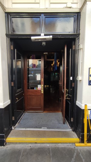 From sidewalk facing venue. Small step before doors. Black doors on top of staircase. Push in centre to open.