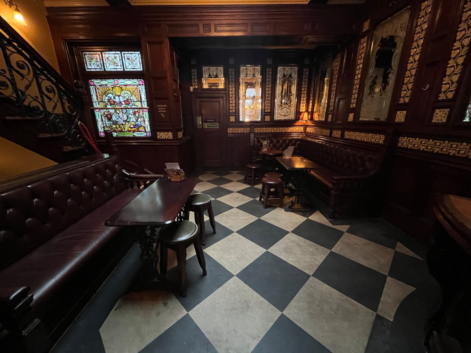 From further along path facing straight. Half booths with stools along left and right walls. Staircase begins to upstairs area at back left.