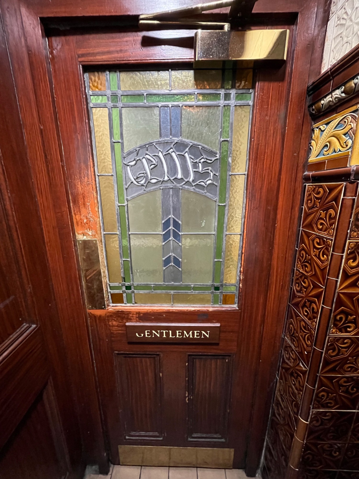 From bottom of stairs facing straight. Men’s bathroom door ahead. Push from left side to open.