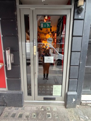 Facing entrance door. Grey framed door. Push silver metal handle on left to open.
