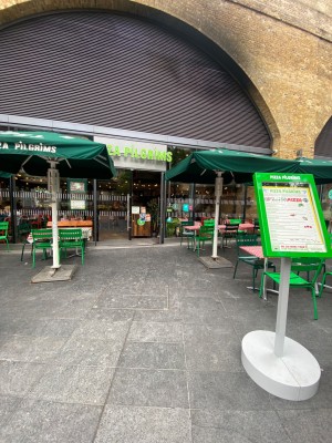 From pavement, facing venue. Menu stand on right. Path ahead to entrance. Tables and chairs along left and right.