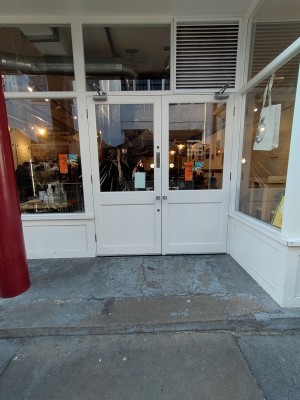 From the street looking forward. There is a double glass door with the handles in the middle. The doors open by pushing. There is a step before the door.