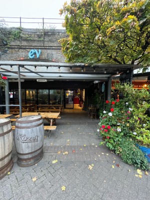 From sidewalk facing venue. Path to entrance ahead. Barrels followed by benches along left. Planters along right.