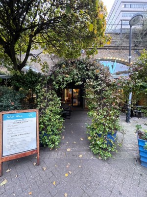 From sidewalk facing venue. A-frame sign on left. Floral archway straight ahead. Path to entrance behind. Seating along left of path.